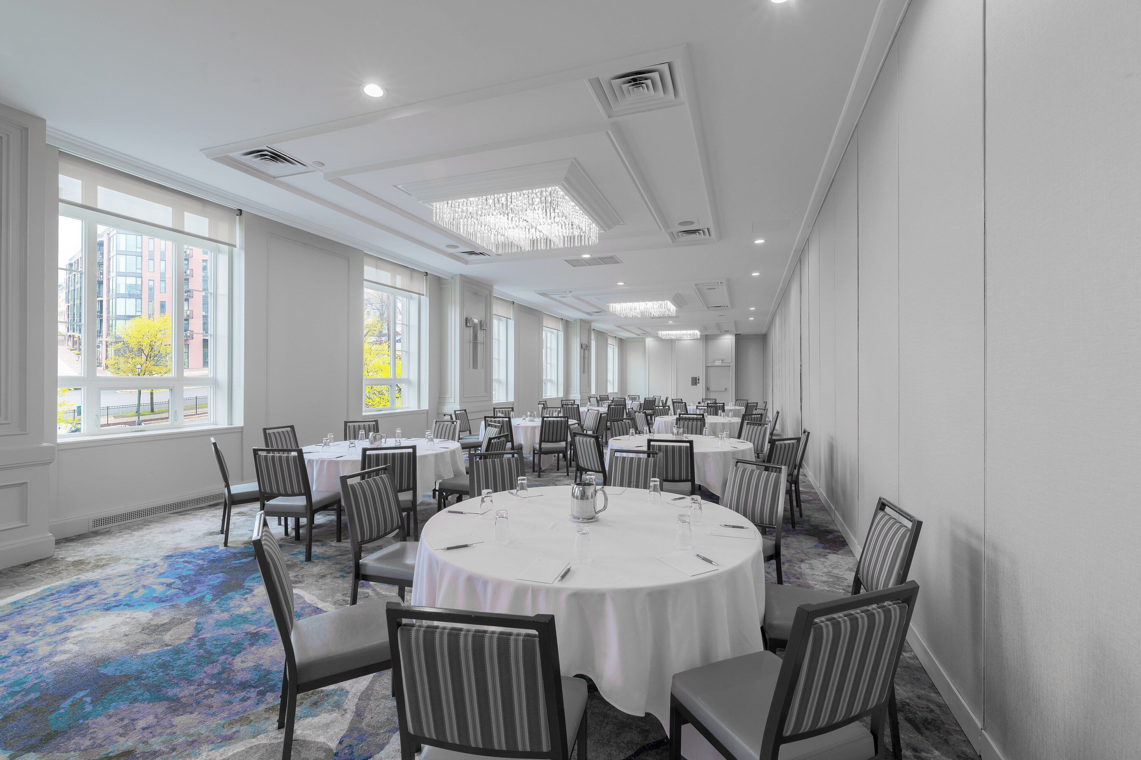 Une salle de conférence dotée de plusieurs tables et chaises, de grandes fenêtres et d’un tapis à motifs.