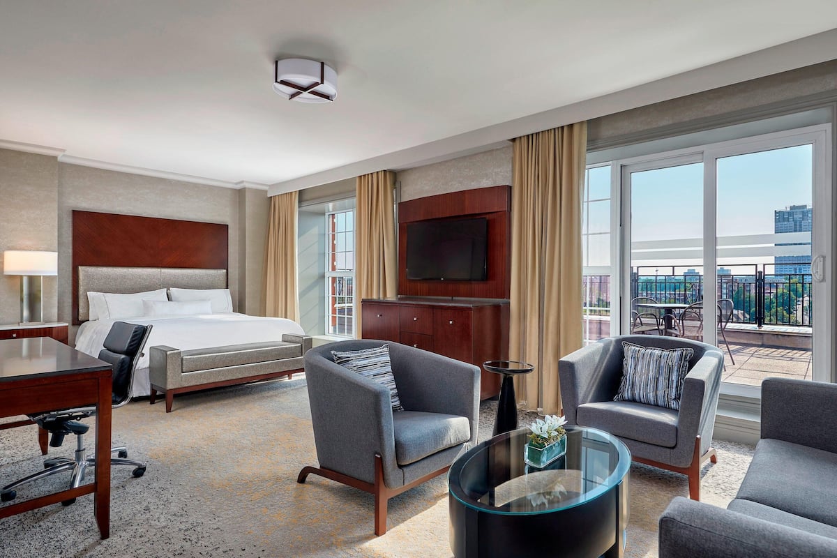 A hotel room with a bed, two armchairs, a desk, a TV, and a balcony with a view of the city.