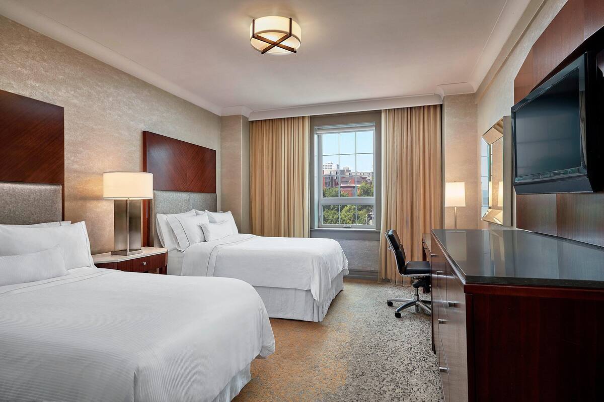 A hotel room with two beds, a desk, a TV, and a window with curtains.