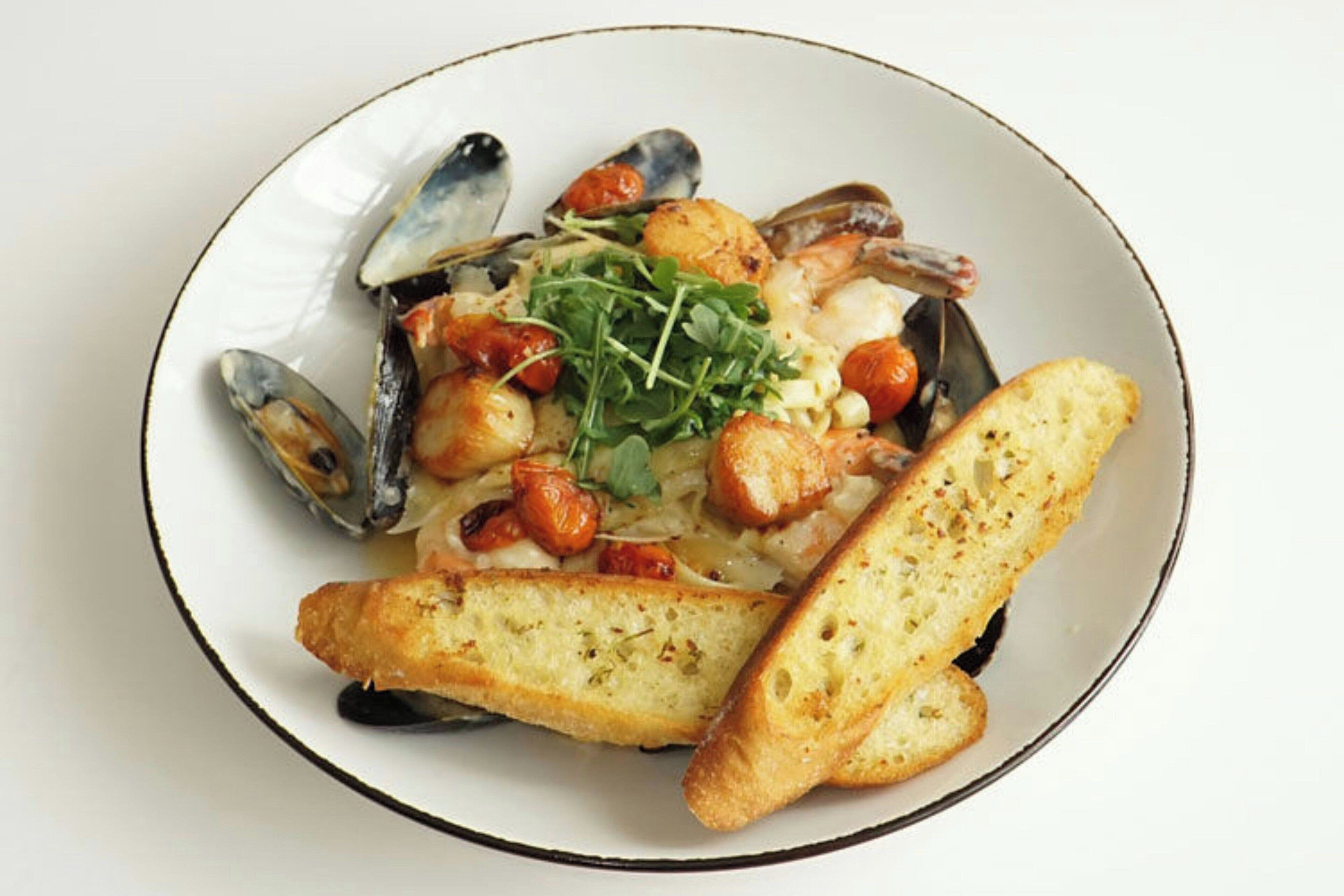 Un plato de mejillones con ajo, tomates cherry y verduras, acompañado de pan tostado.