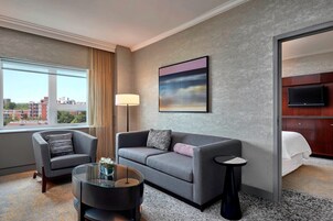 Habitación de hotel con un sofá gris, un sillón más pequeño, una mesa de centro de vidrio y un televisor montado en la pared.