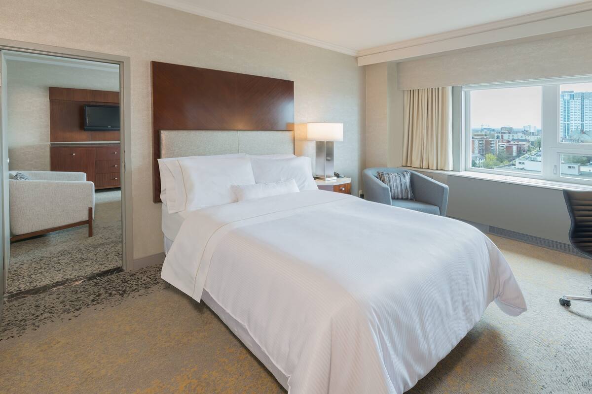 A hotel room with a large bed, a television on a wooden cabinet, a desk with a lamp, and a view of the city through the window.