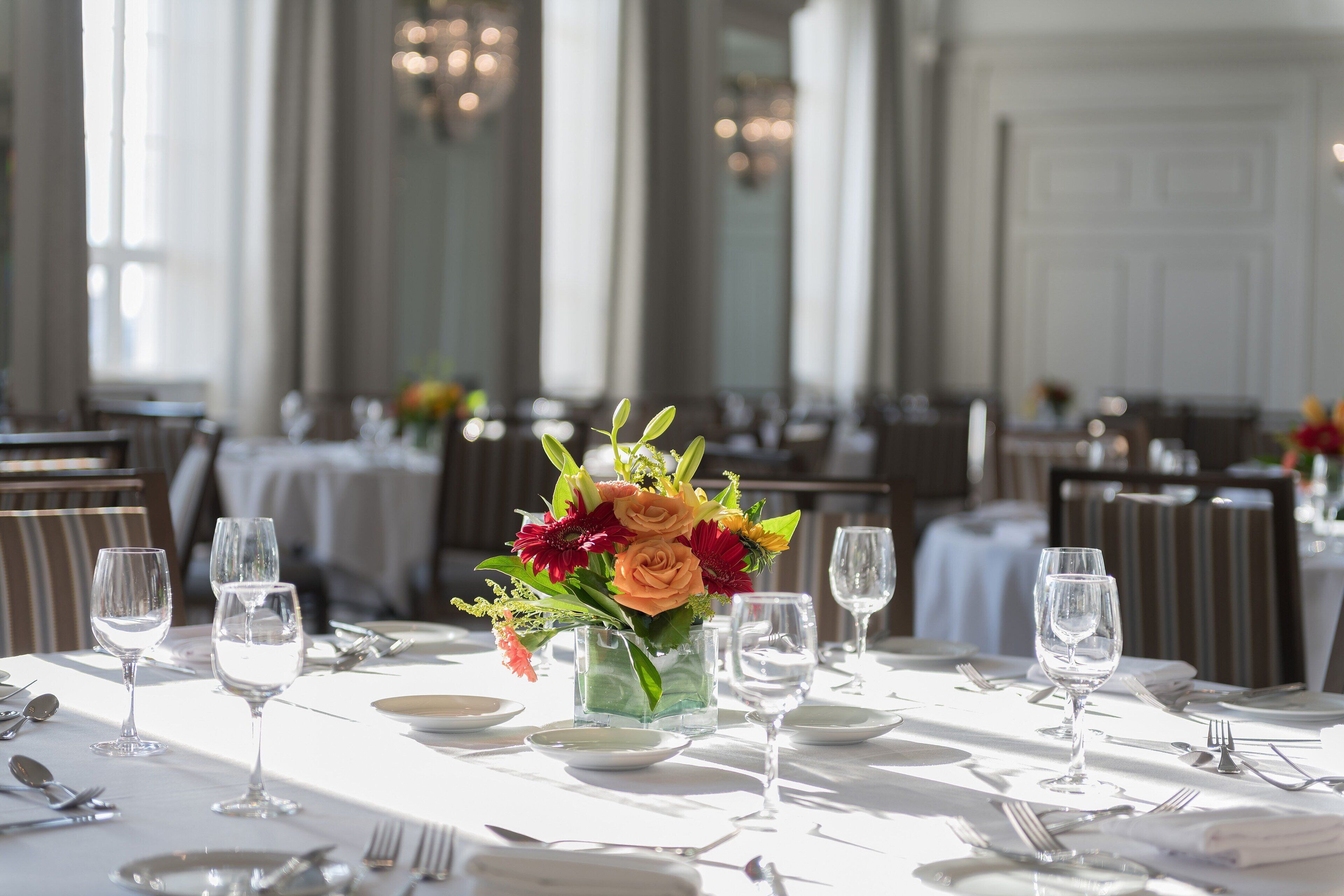 A dining table set with white tablecloths, glassware, and a centerpiece of flowers in a glass vase.