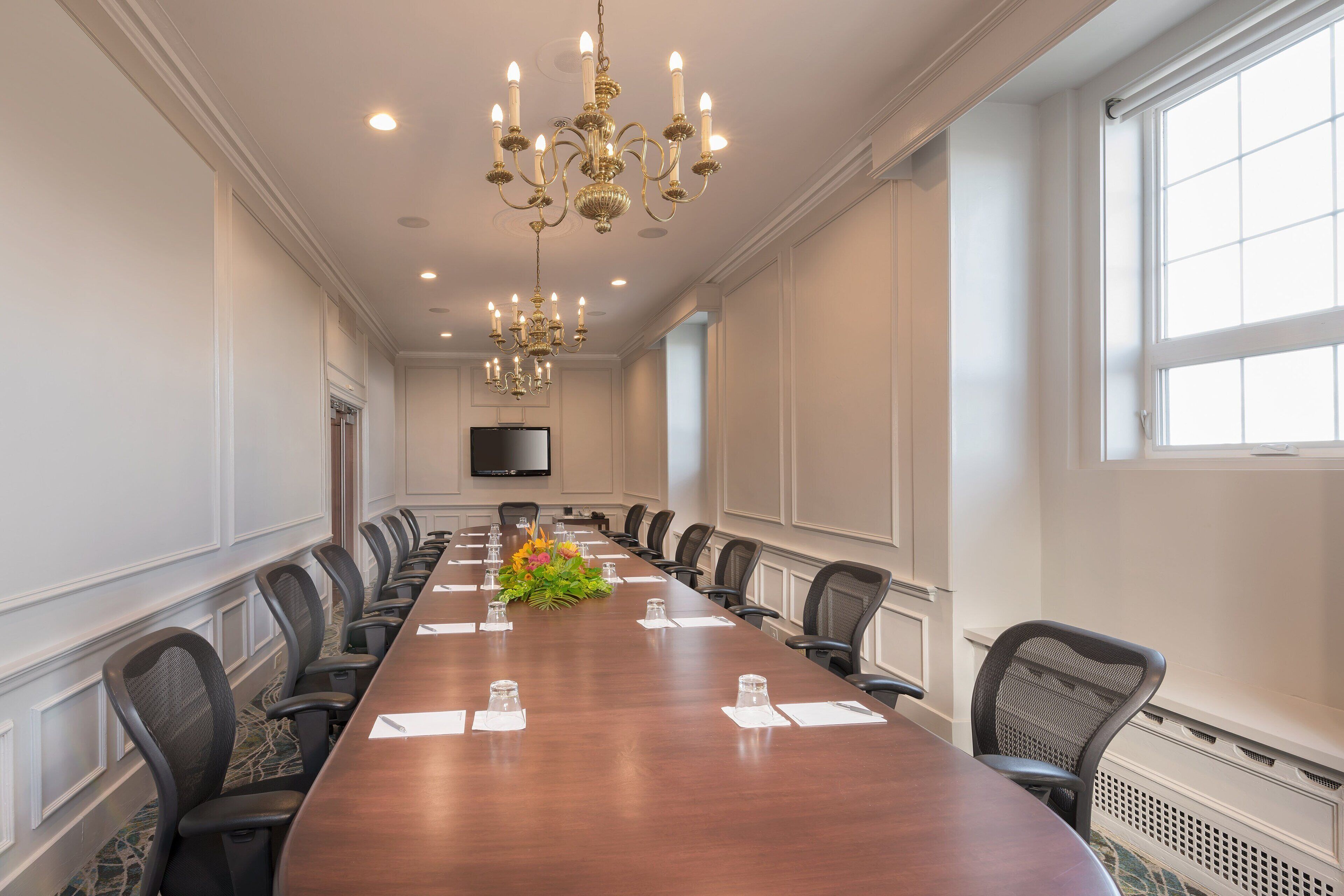 A conference room with a long table, chairs, a chandelier, and a TV on the wall.