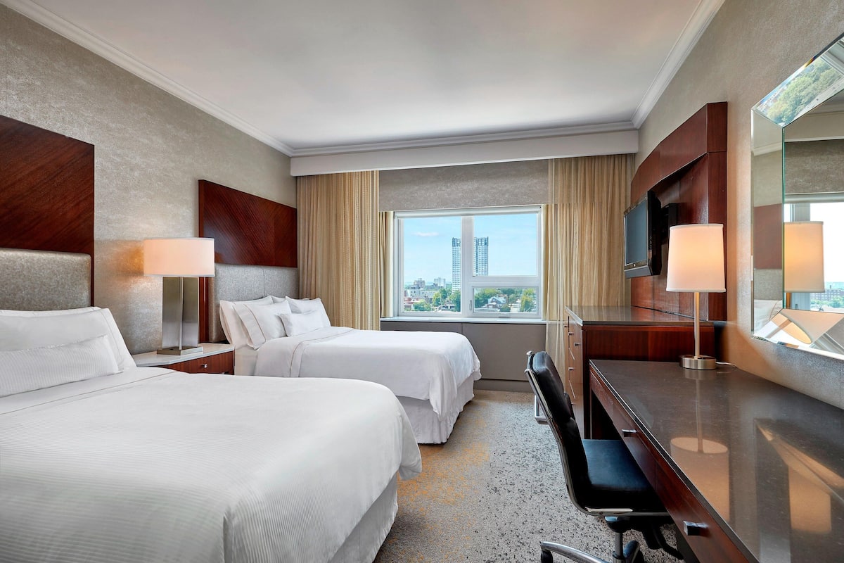 A hotel room with two beds, a desk, a chair, a TV, and a large window with a city view.