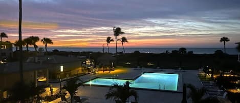 A swimming pool area with lounge chairs and palm trees, surrounded by buildings and a sunset view.