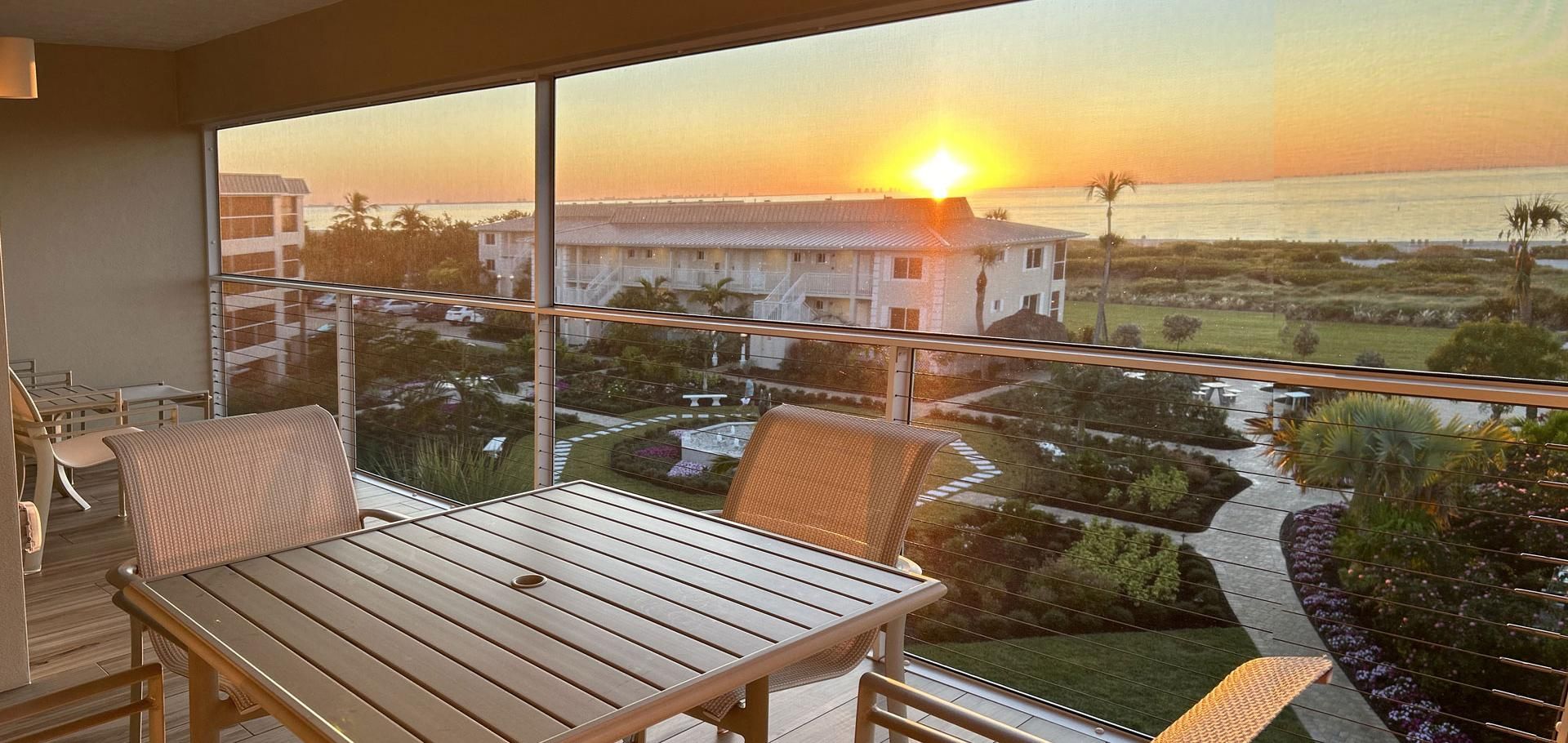 A balcony with a wooden table and chairs, overlooking a residential area and the ocean at sunset.