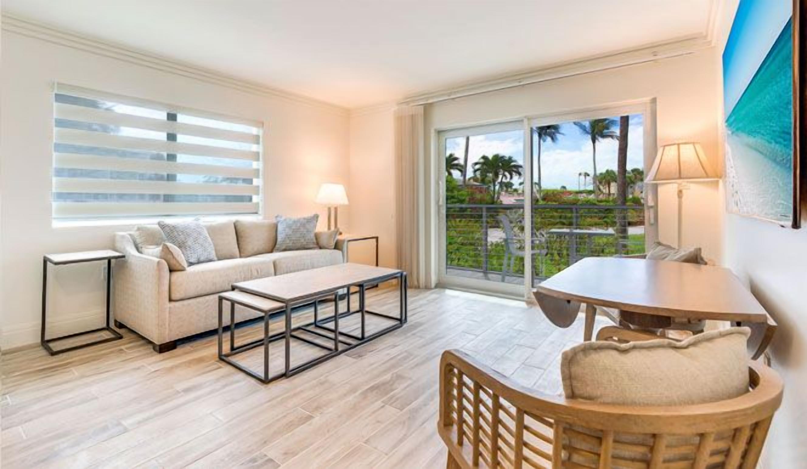 A hotel room with a sofa, a coffee table, a dining table, and a chair. There is a window with blinds, a door leading to a balcony, and a painting on the wall.