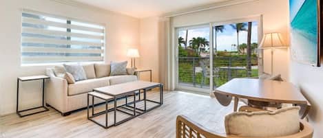 A hotel room with a sofa, a coffee table, a dining table, and a chair. There is a window with blinds, a door leading to a balcony, and a painting on the wall.