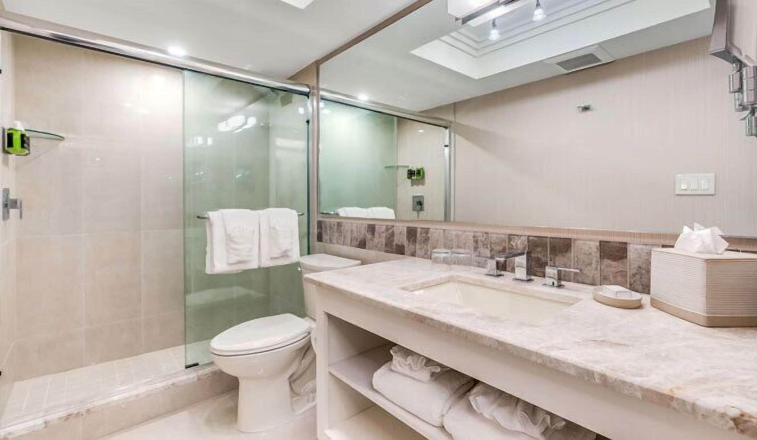 Une salle de bain moderne avec une douche à l’italienne en verre, des toilettes blanches, un meuble-lavabo en marbre avec un évier et un grand miroir.