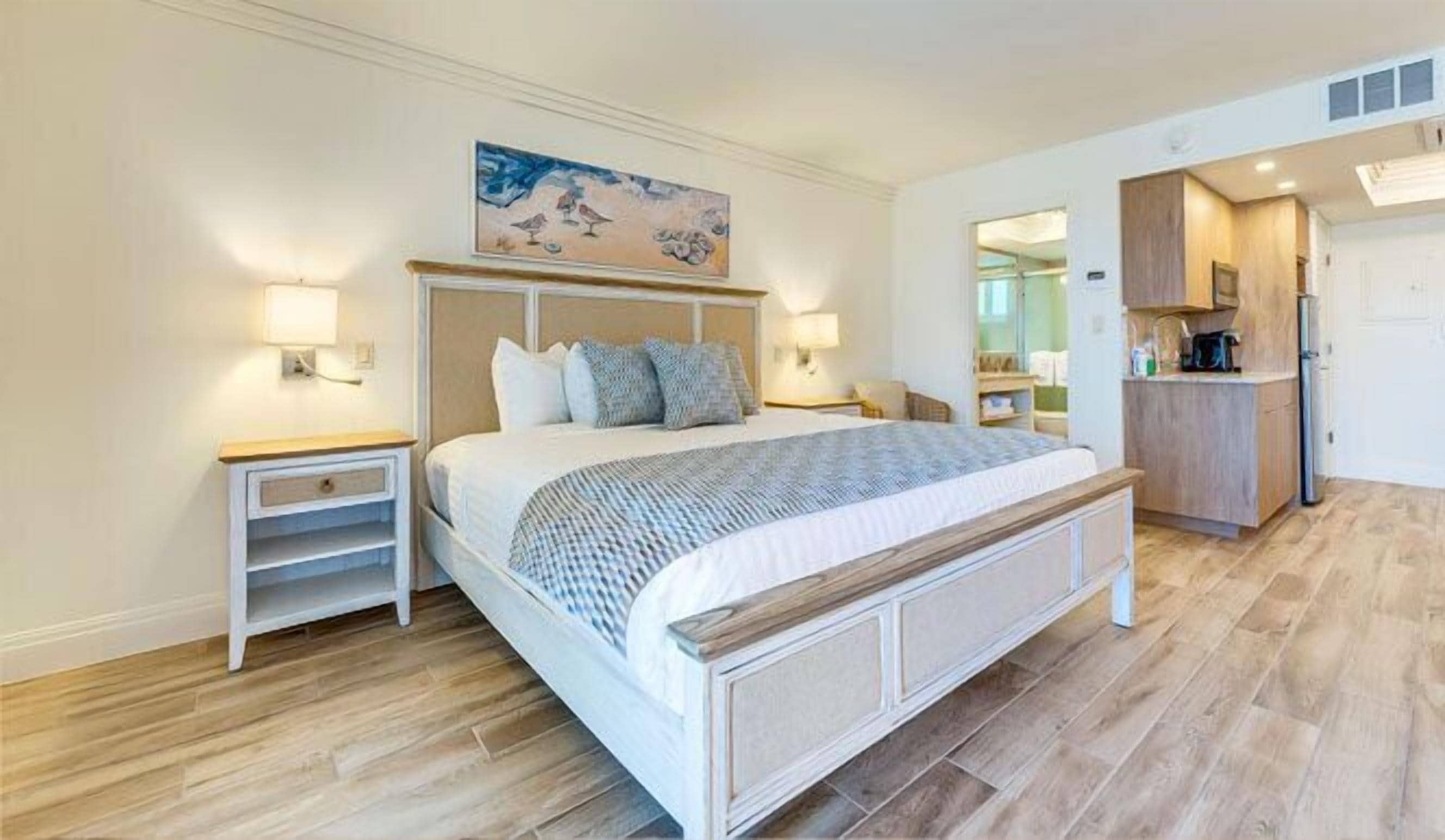 A bedroom with a large bed, bedside tables, a painting above the bed, and a kitchenette area.