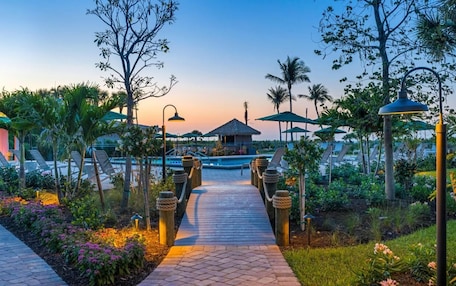 A pathway leading to a pool area with lounge chairs and umbrellas, surrounded by tropical plants and a thatched-roof structure.