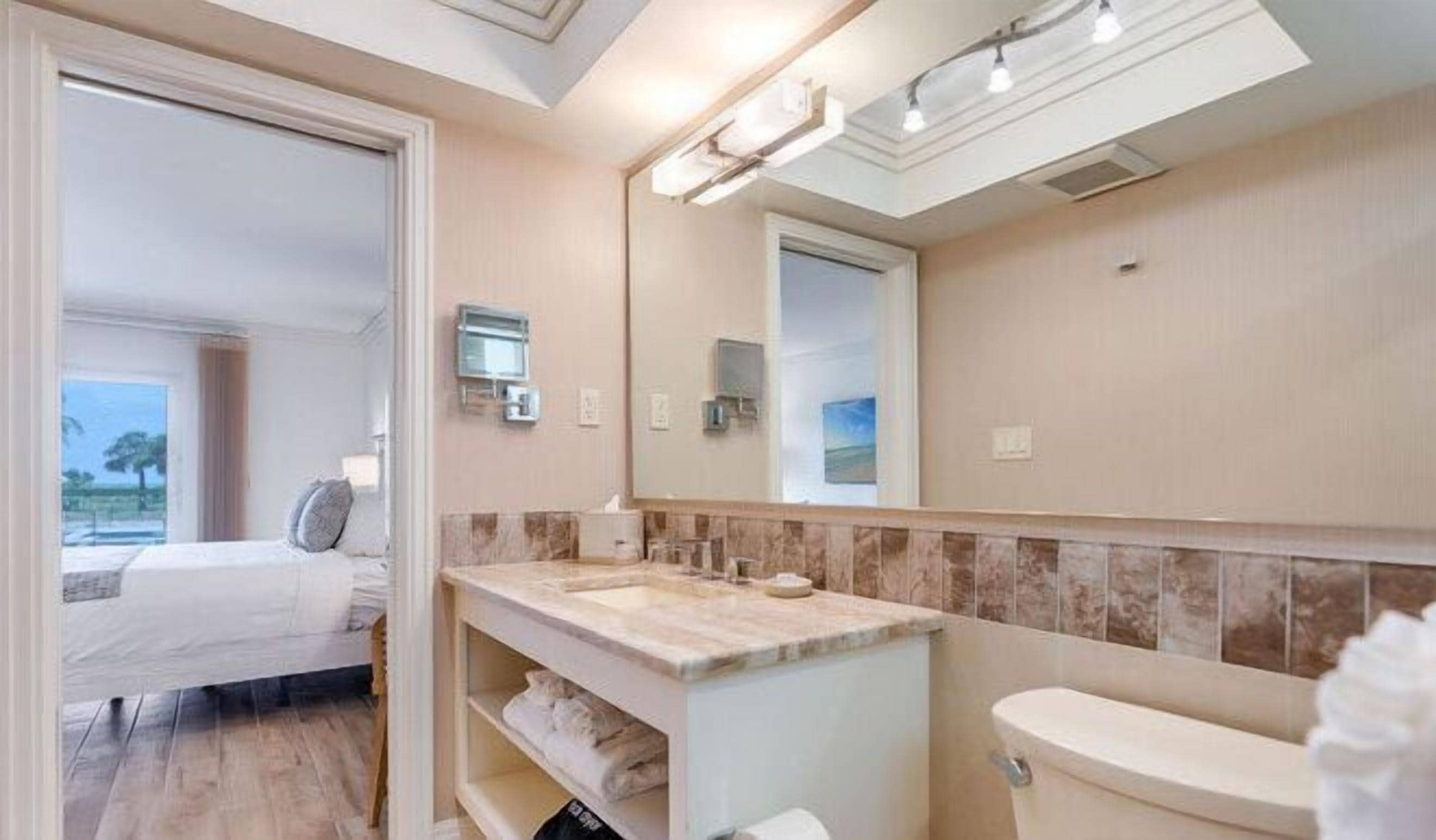 Un baño moderno con un espejo grande, un lavamanos y un inodoro. Se ve una cama en la habitación contigua.