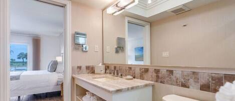 Un baño moderno con un espejo grande, un lavamanos y un inodoro. Se ve una cama en la habitación contigua.