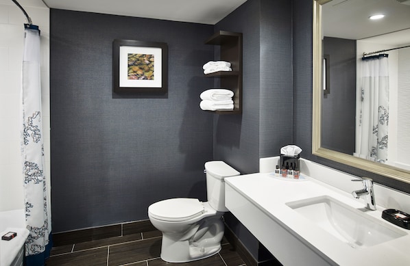 A modern bathroom with a white toilet, a sink, and a shower curtain. There is a framed picture on the wall and a shelf with folded towels.