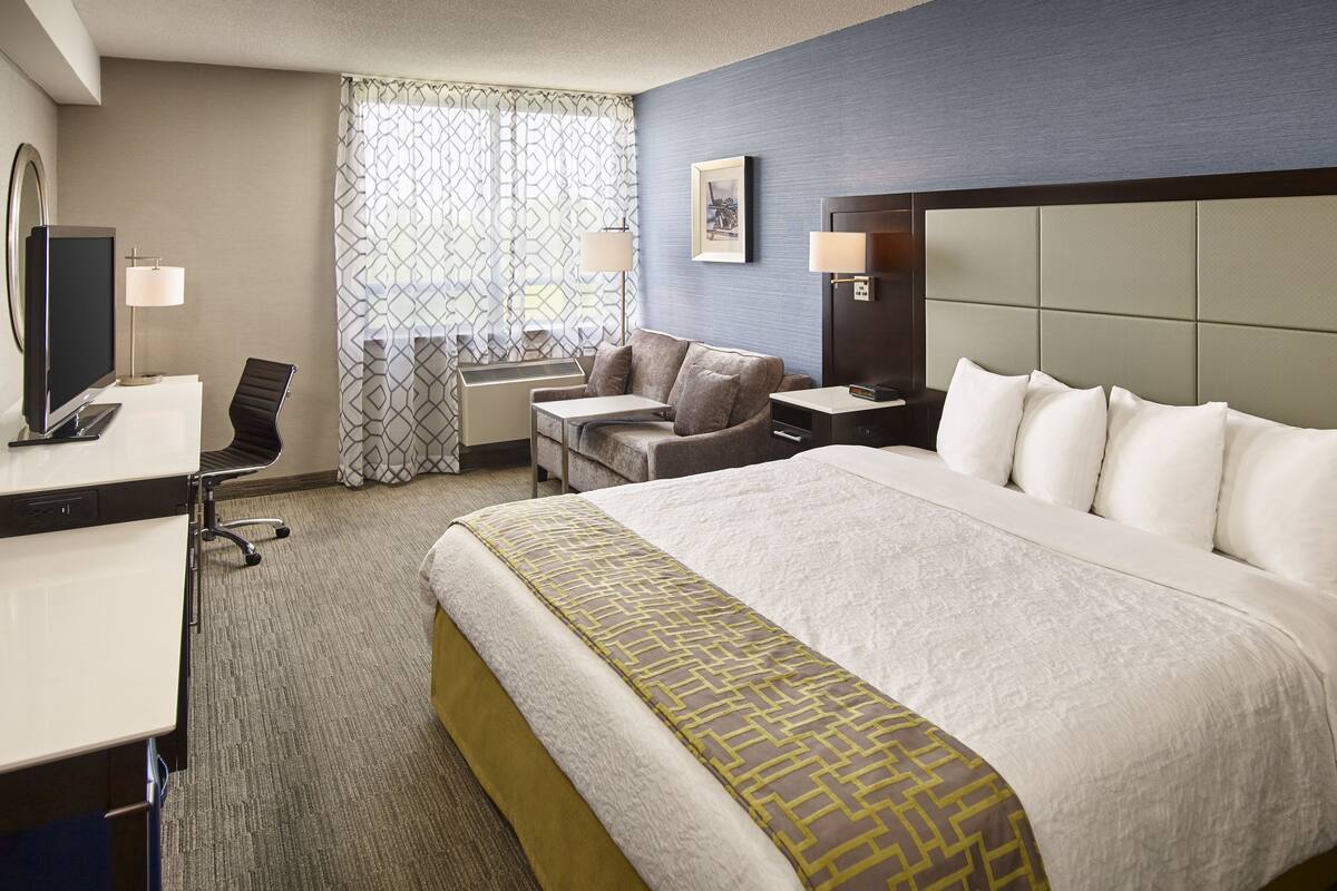A hotel room with a large bed, a desk with a computer, a chair, a small table, and a window with patterned curtains.