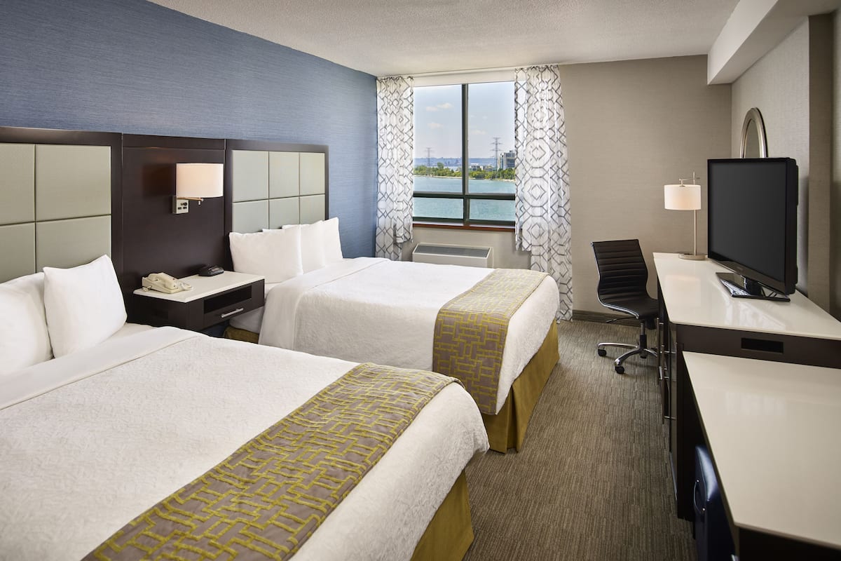 A hotel room with two beds, a desk, a TV, and a window with a view of the city.
