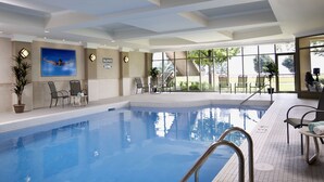 An indoor swimming pool with clear water, surrounded by tiled walls and a ceiling with recessed lighting. There are chairs and a potted plant near the pool, and a large window with a view of greenery outside. - Waterfront Hotel Downtown Burlington (Burlington)