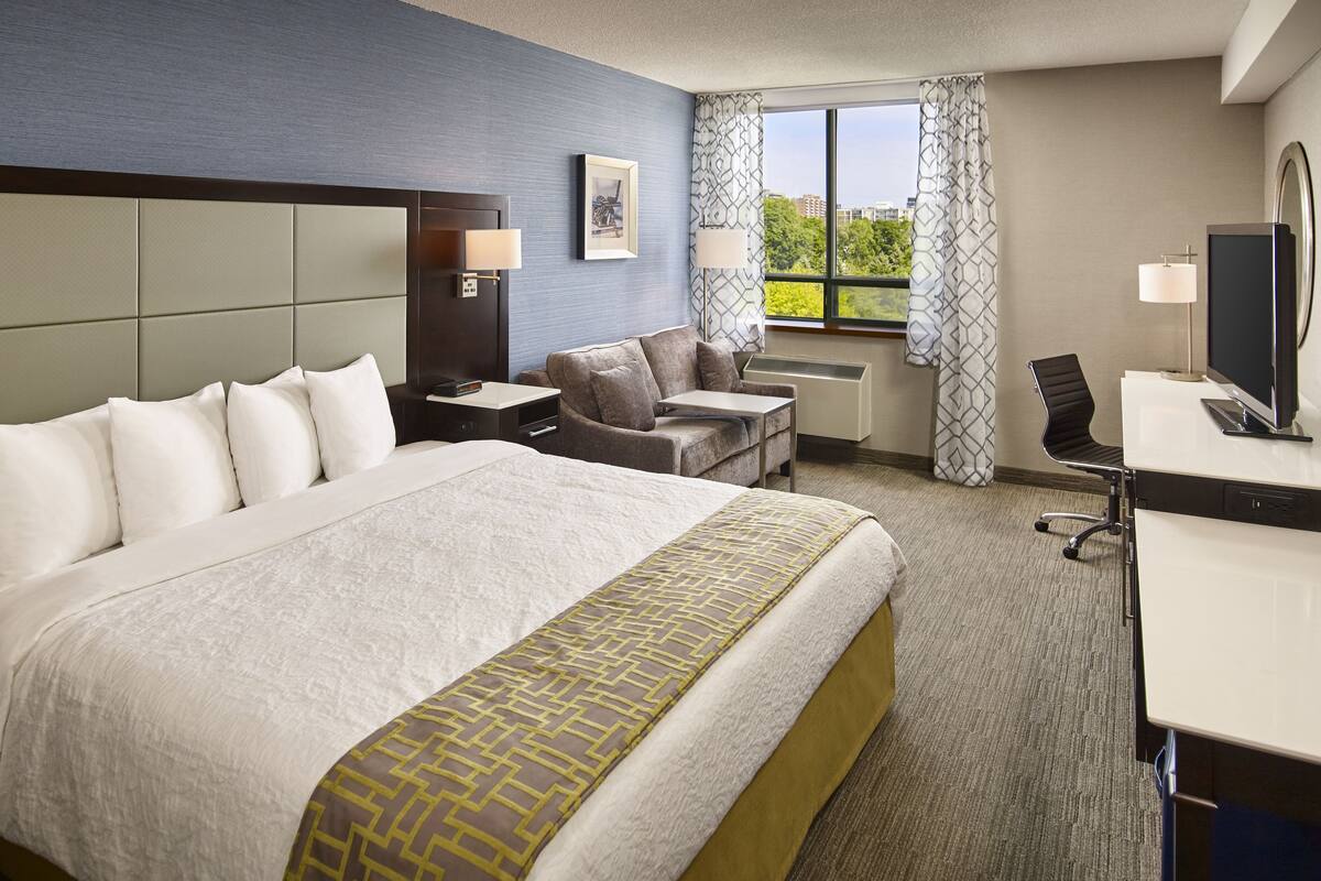 A hotel room with a large bed, a sofa, a desk with a computer, and a window with curtains.