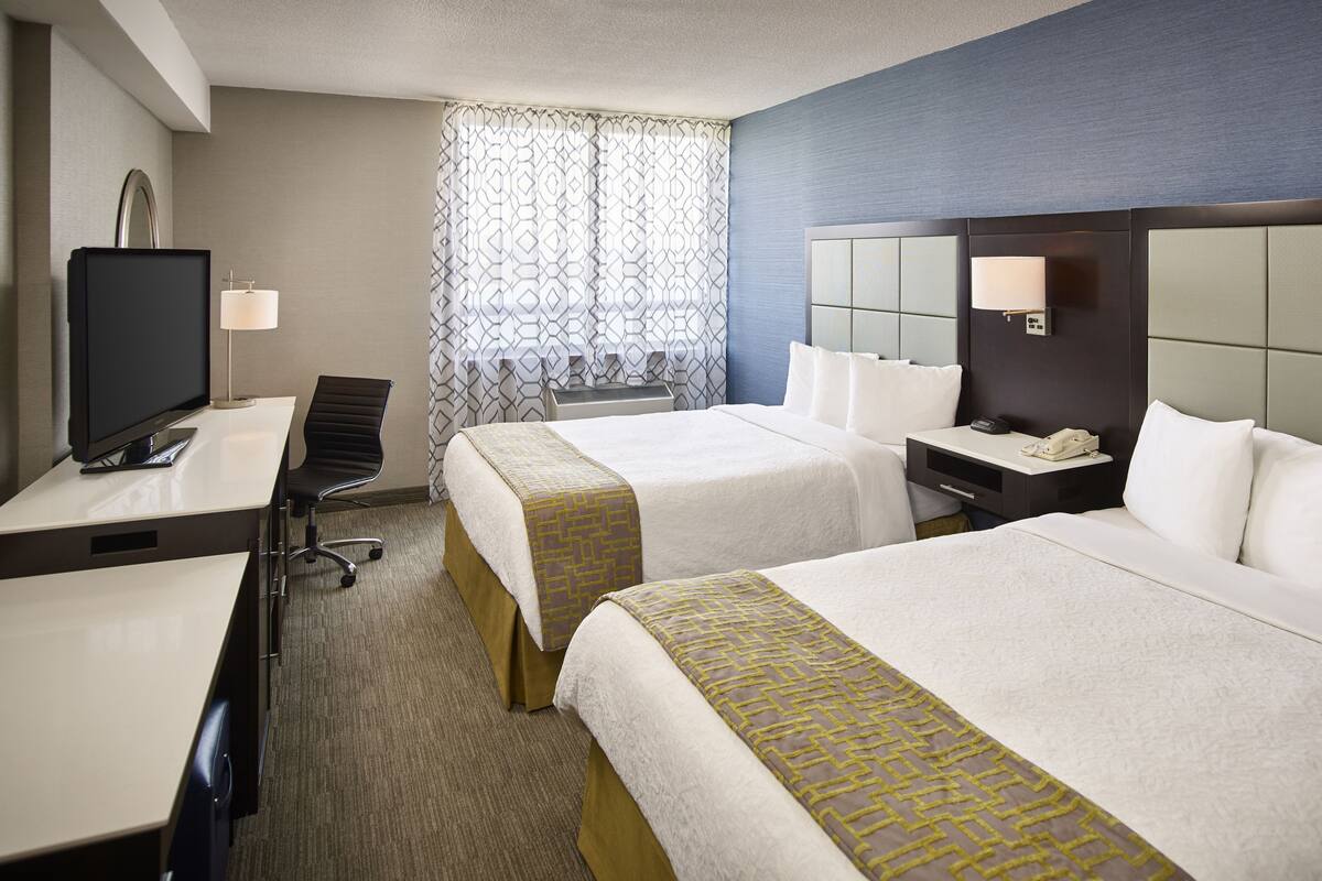 A hotel room with two beds, a desk with a TV, a chair, a nightstand with a lamp, and a window with patterned curtains.
