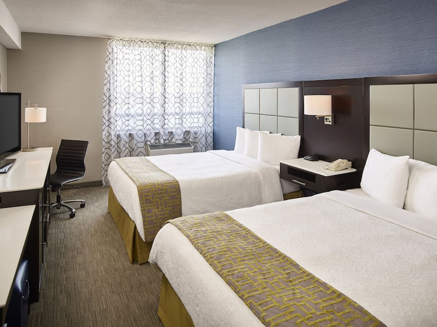 Une chambre d’hôtel avec deux lits, un bureau avec une télévision, une chaise, une table de chevet avec une lampe et une fenêtre avec des rideaux à motifs.