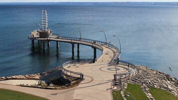 A waterfront promenade with a curved bridge, a circular viewing area, and a lighthouse in the background.