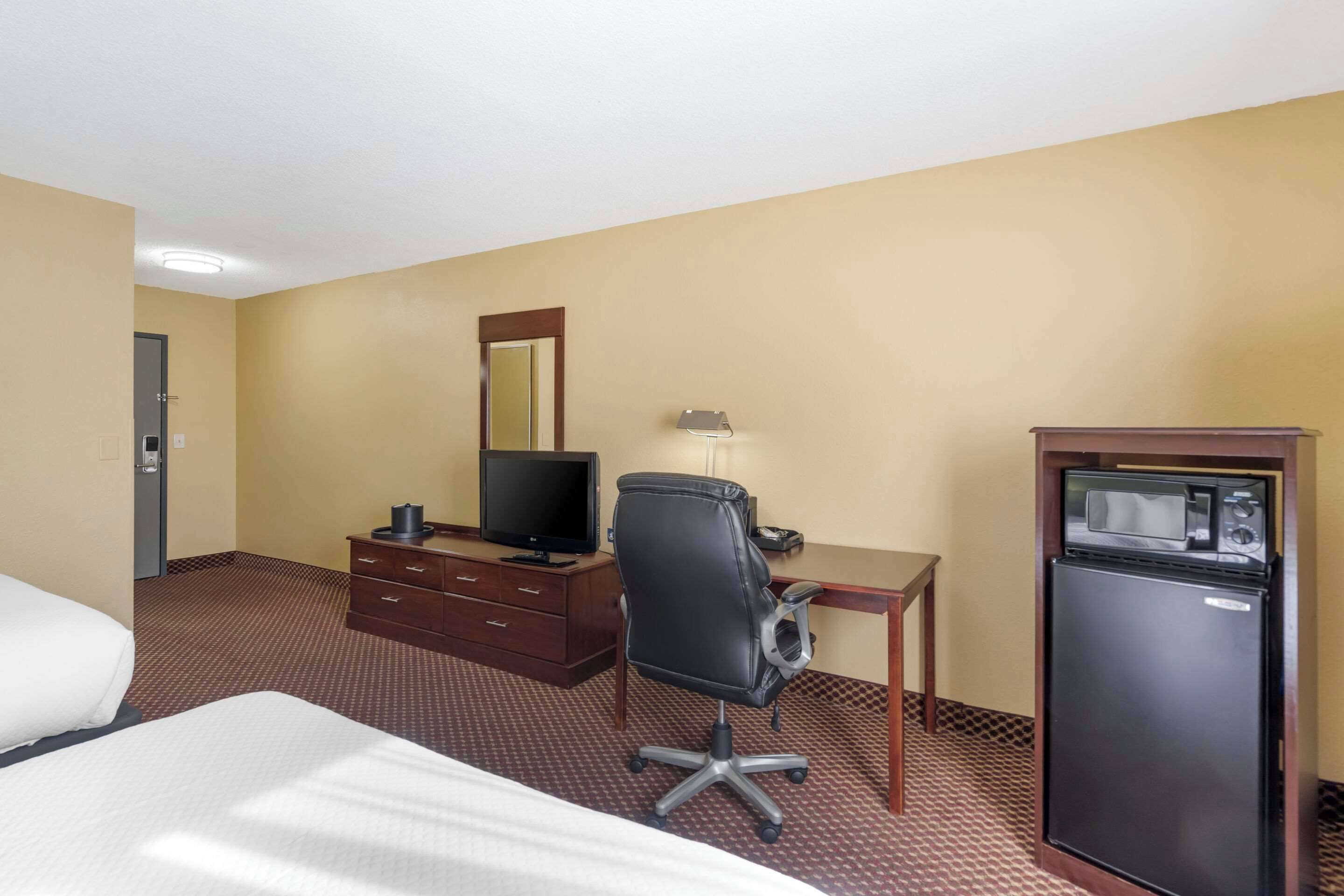 A hotel room with a bed, a desk with a computer, a chair, a small refrigerator, and a mirror on the wall.