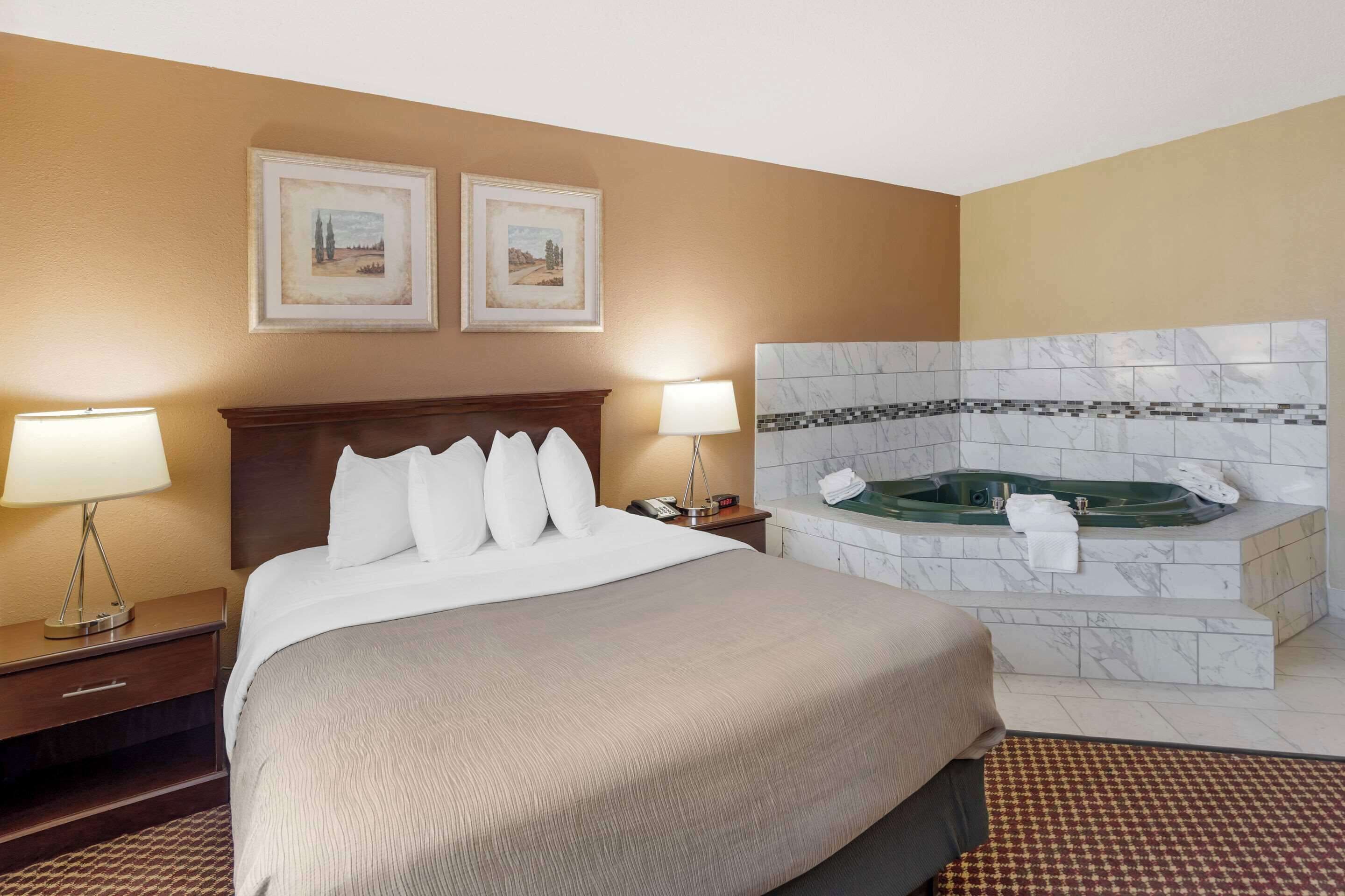 A hotel room with a large bed, two bedside lamps, a nightstand, a framed picture, and a marble-tiled bathtub.