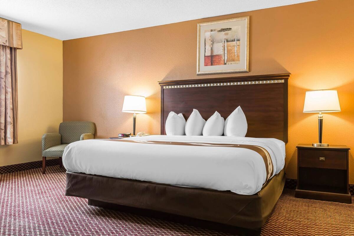 A hotel room with a large bed, two bedside tables with lamps, a chair, a framed artwork on the wall, and a patterned carpet.