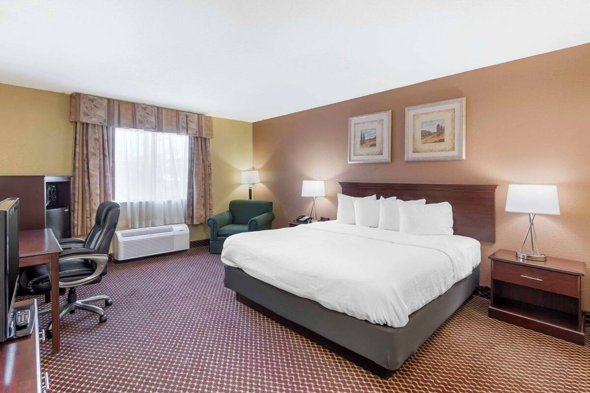 A hotel room with a large bed, a desk with a computer, a chair, two lamps, and a heater.