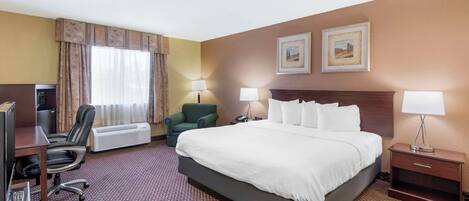 A hotel room with a large bed, a desk with a computer, a chair, two lamps, and a heater.