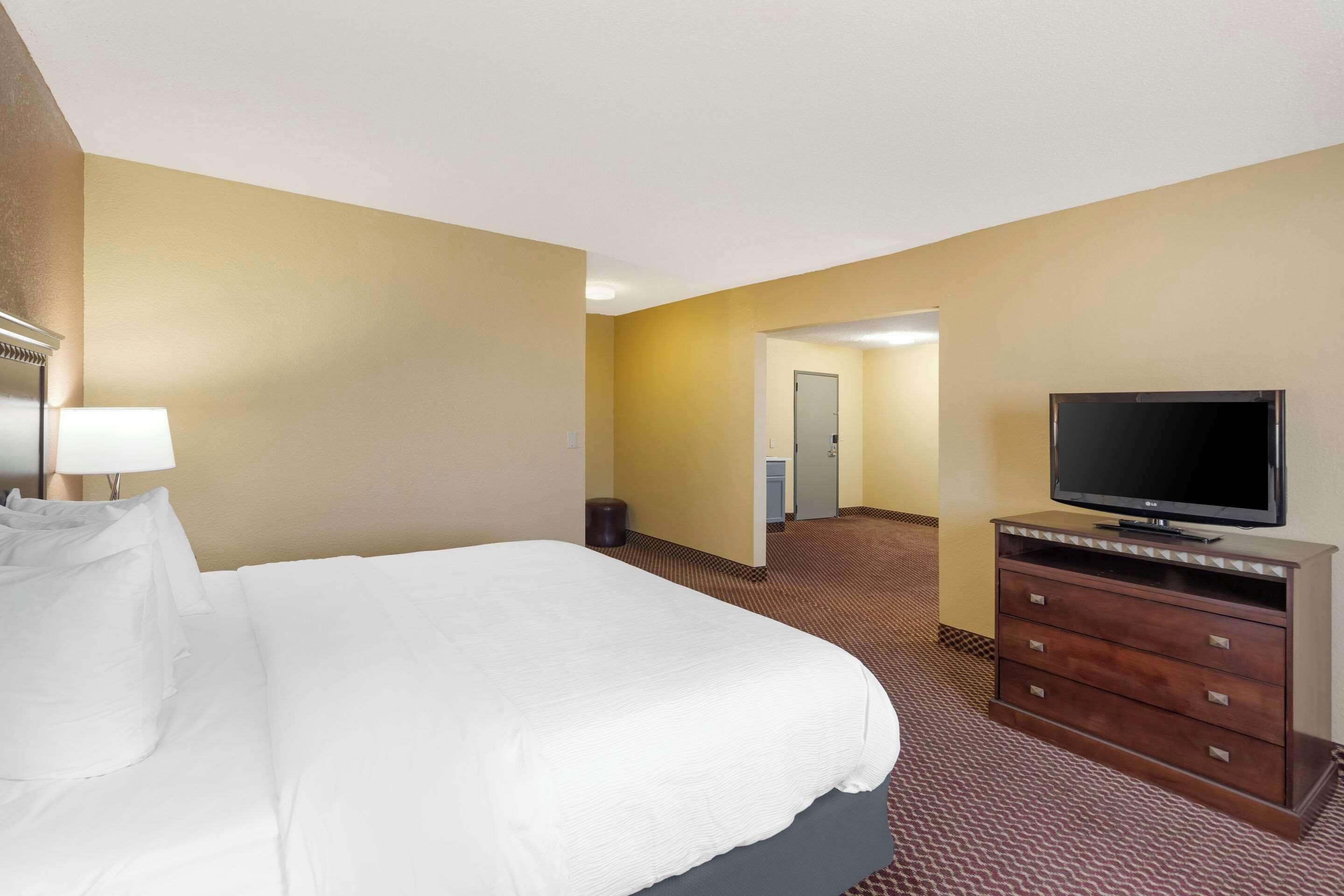 Quarto de hotel com uma cama grande, uma televisão sobre uma cômoda de madeira e piso coberto por carpete.