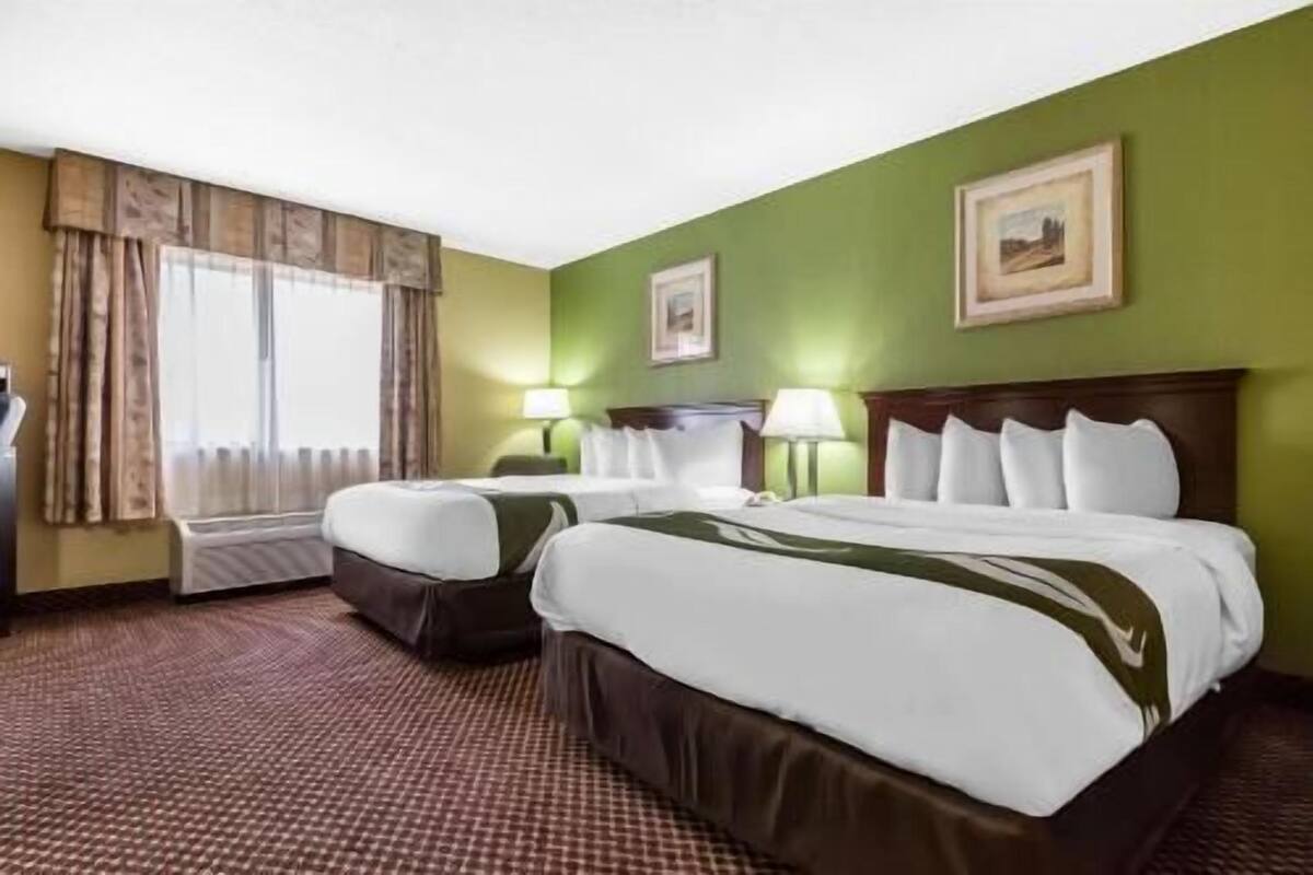 A hotel room with two beds, a desk, a chair, a window with curtains, and two wall-mounted lamps.