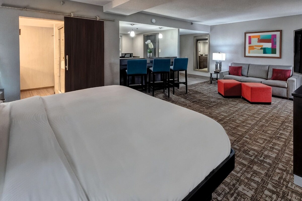 A hotel room with a large bed, a desk with chairs, a sofa, and a colorful abstract painting on the wall.