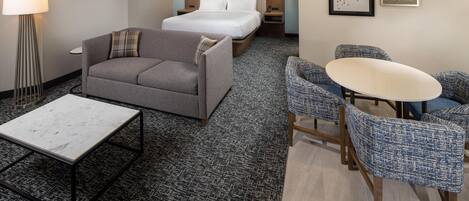 Una habitación de hotel con cama, sofá, una mesita y una zona de estar con sillones.