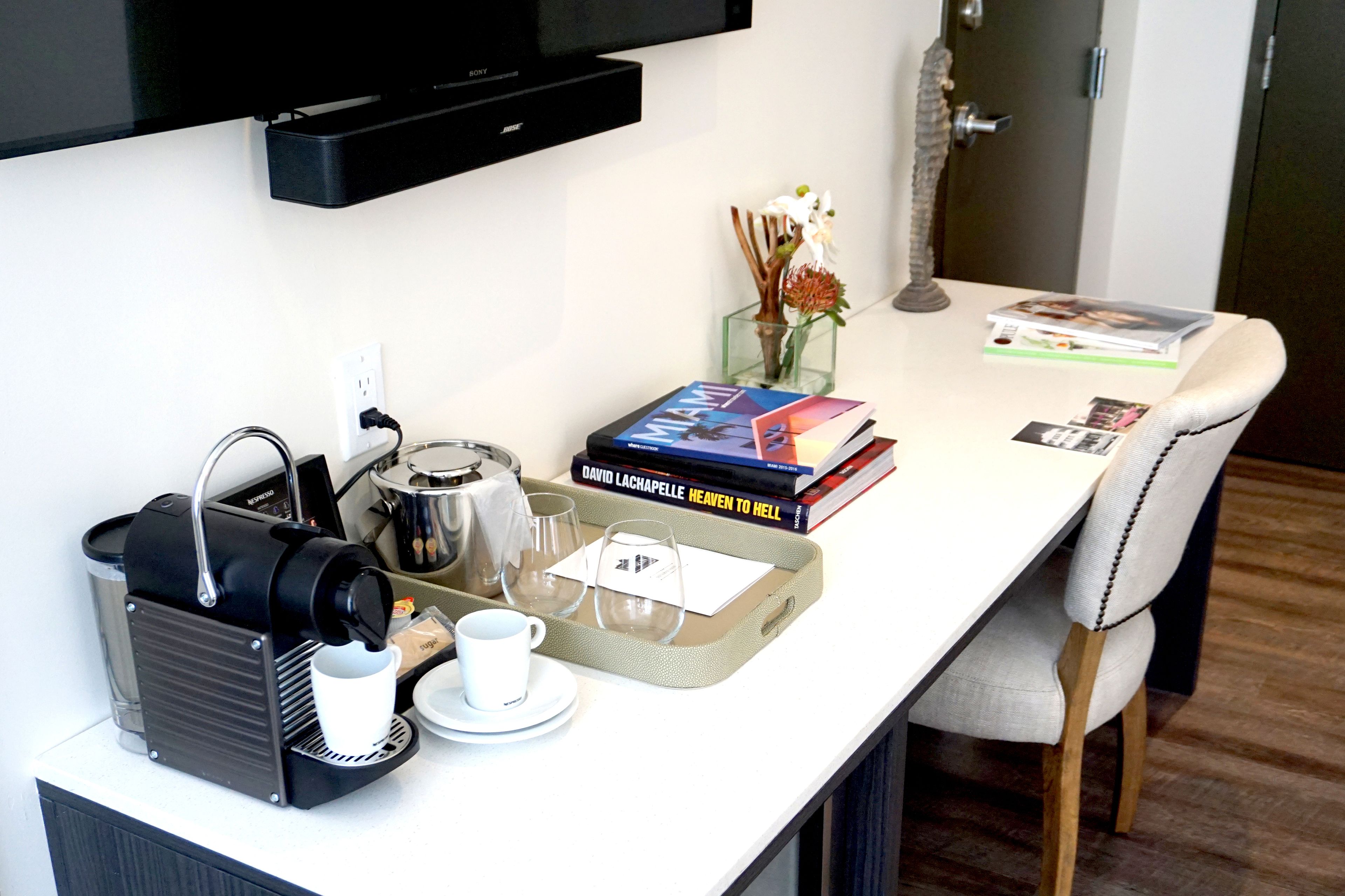 Uma mesa de pequeno-almoço bem arrumada, com uma máquina de café, um tabuleiro com chávena e pires, livros e um vaso decorativo.