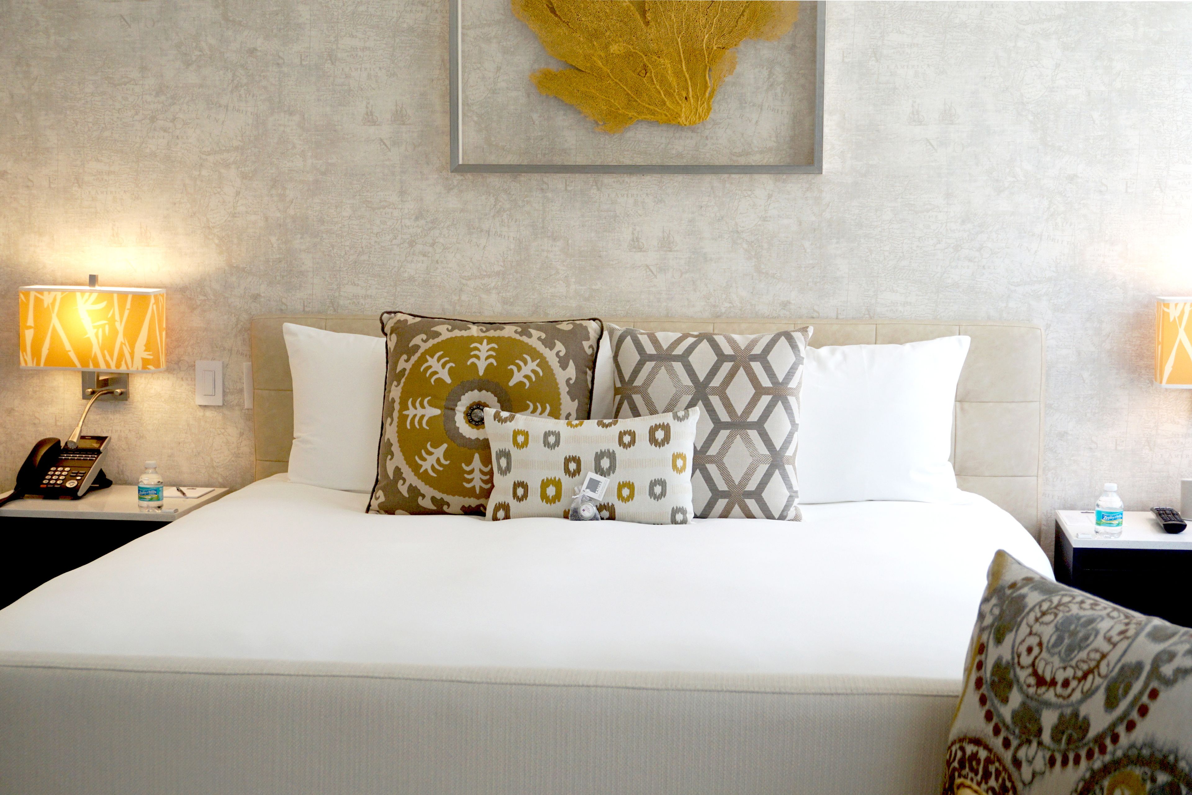 A hotel room with a bed, two bedside tables, a phone, a water bottle, and a framed artwork on the wall.