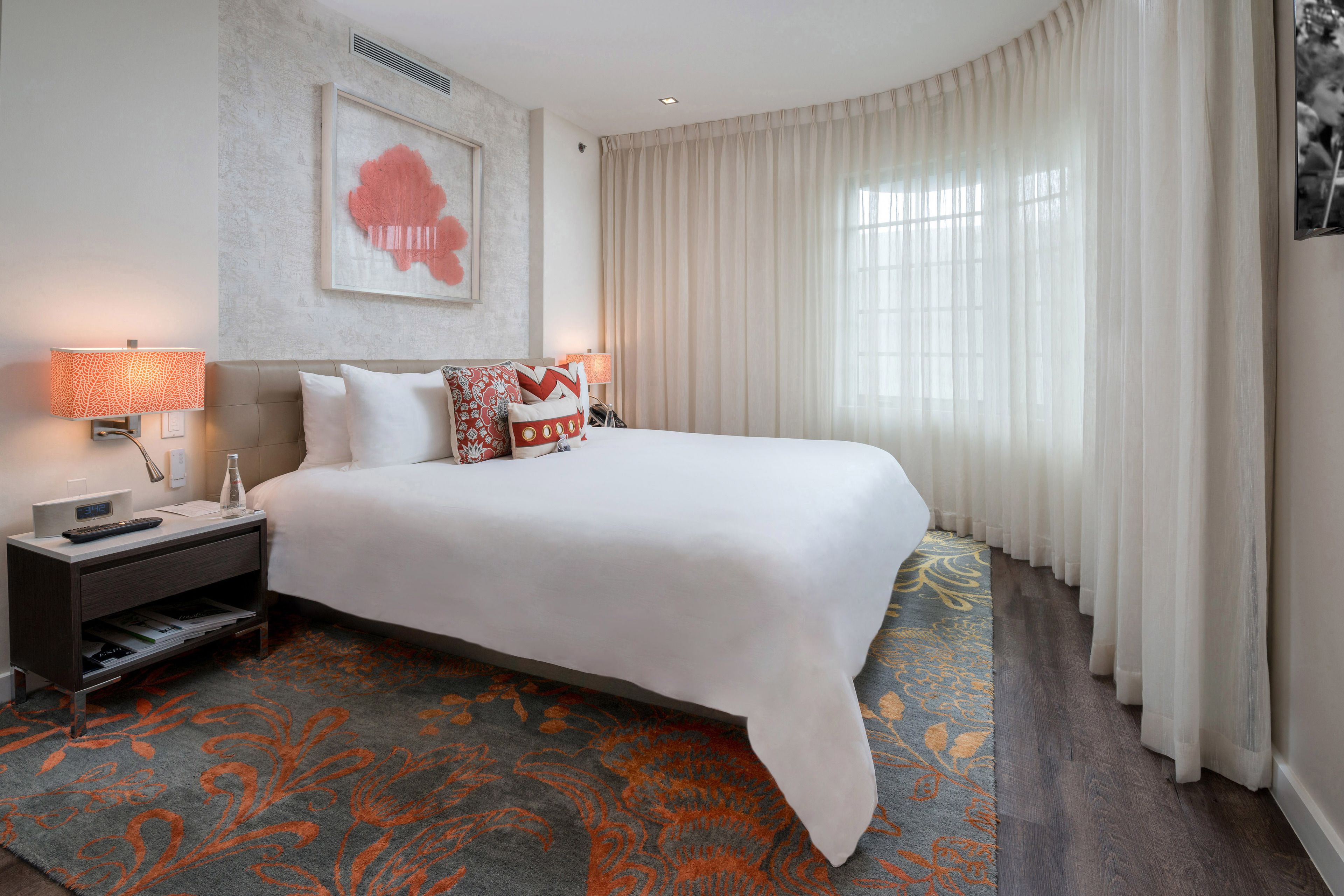 A hotel room with a large bed, a nightstand with a lamp, a framed artwork on the wall, and a patterned carpet.