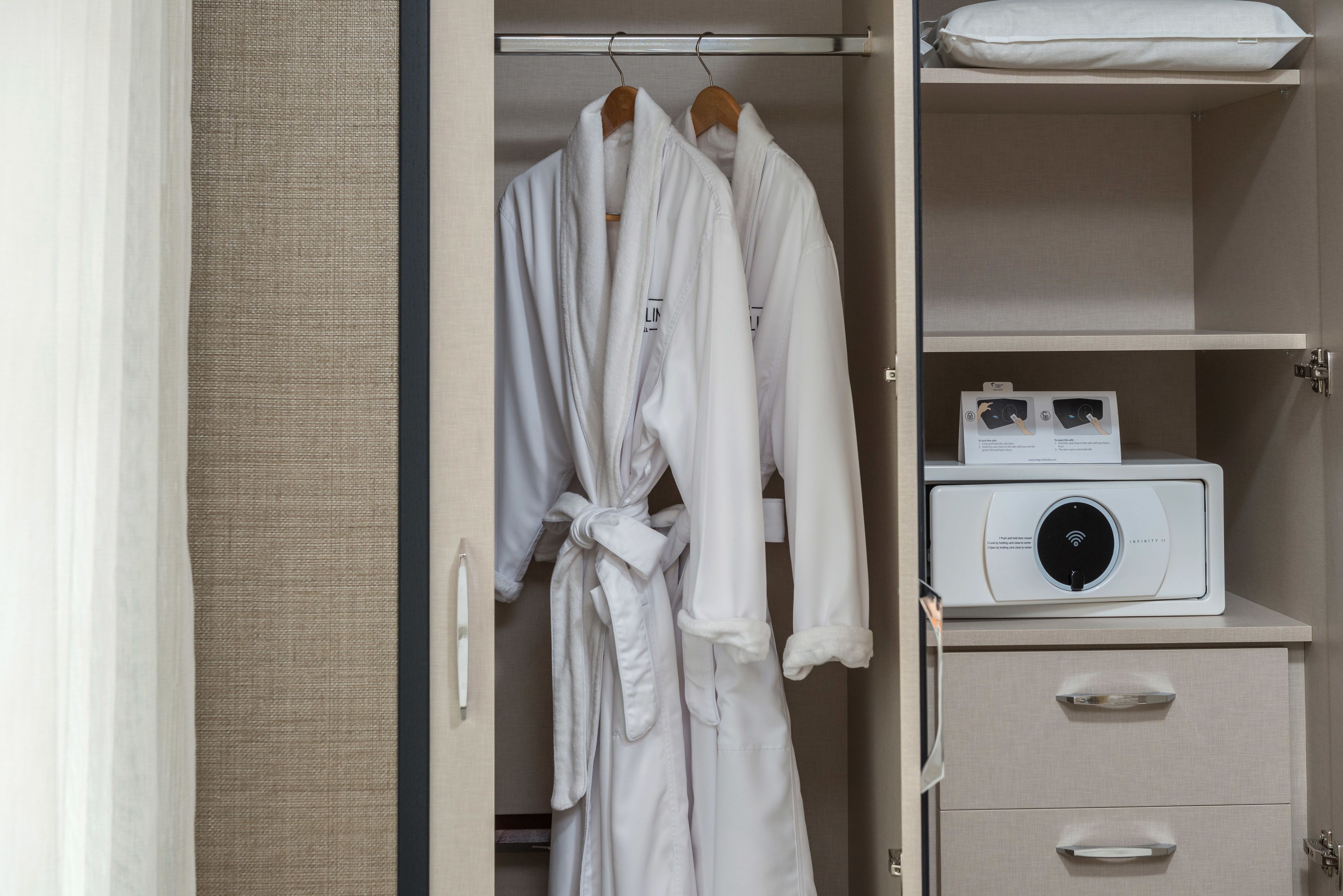 Camera d'albergo con cabina armadio contenente due accappatoi bianchi appesi a un appendiabiti in legno, un comodino bianco con un dispositivo bianco e una parete beige.