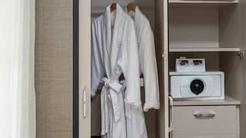 Camera d'albergo con cabina armadio contenente due accappatoi bianchi appesi a un appendiabiti in legno, un comodino bianco con un dispositivo bianco e una parete beige.