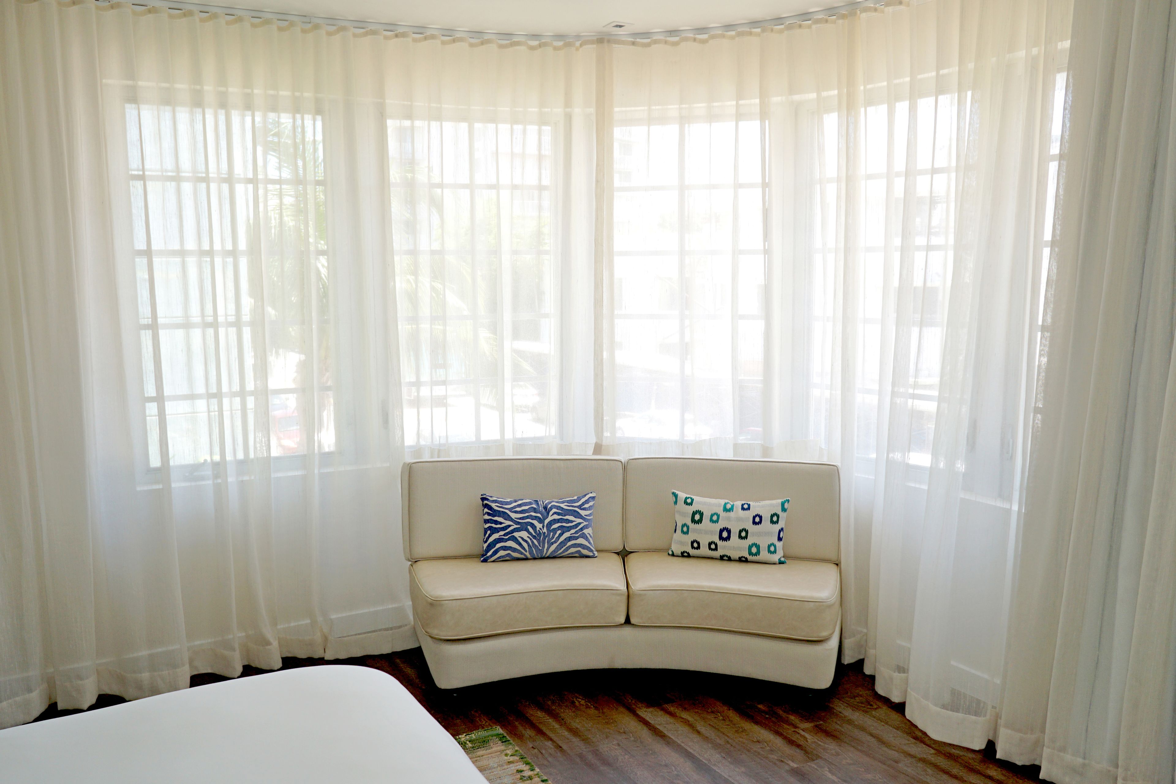 A room with a curved beige sofa, two patterned cushions, and sheer white curtains.
