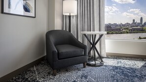 Habitación de hotel moderna con vistas a la ciudad, un sillón oscuro, una mesa redonda y un amplio ventanal con cortinas.
