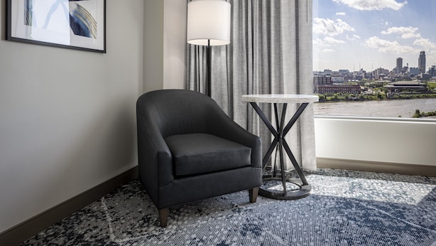 Une chambre d’hôtel moderne avec vue sur la ville, un fauteuil sombre, une table ronde et une grande fenêtre avec des rideaux.