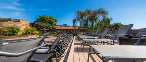 A sunlit outdoor seating area with rows of metal chairs and tables, surrounded by lush greenery and a clear blue sky.