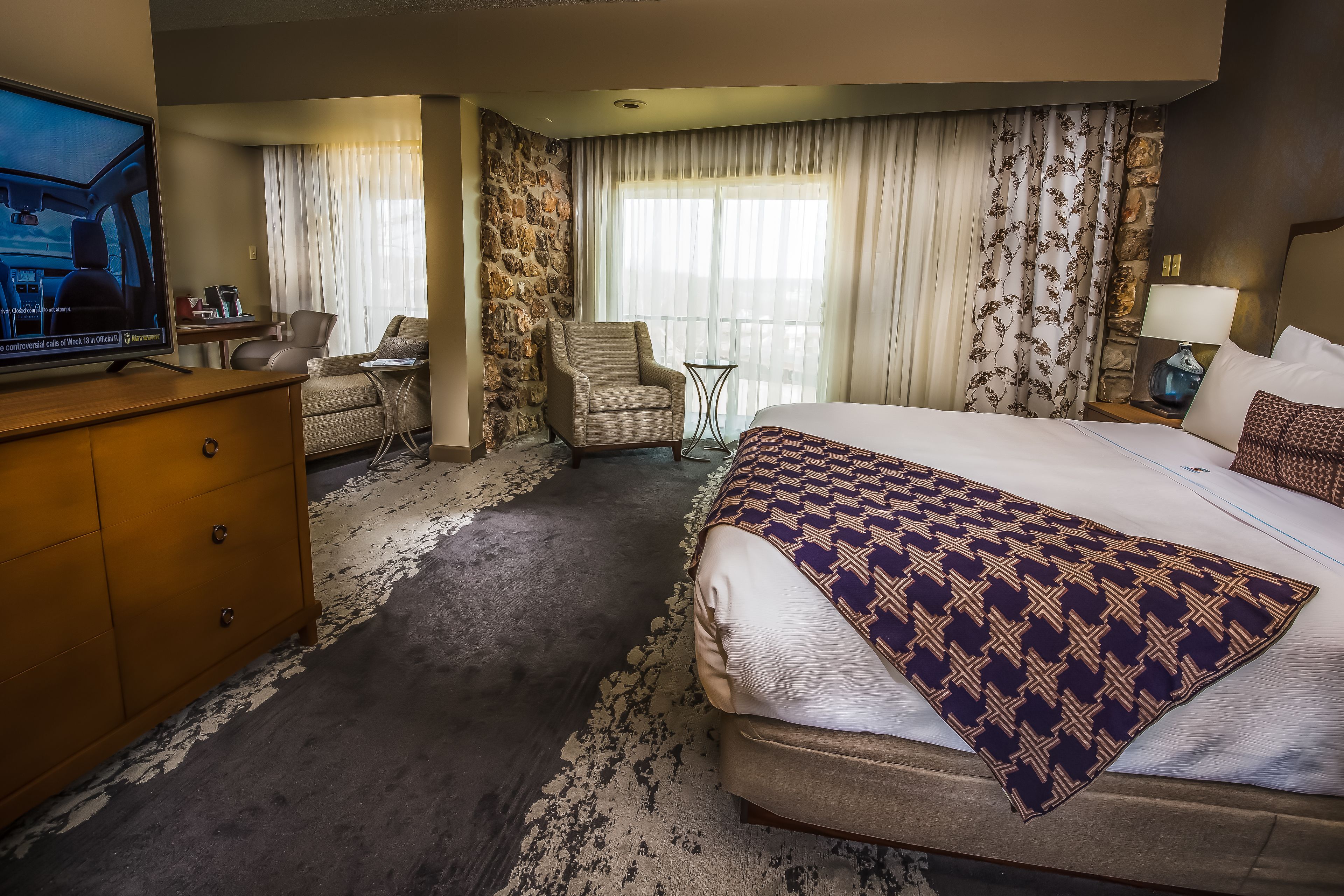 A hotel room with a large bed, a desk with a chair, a television, and a stone accent wall.