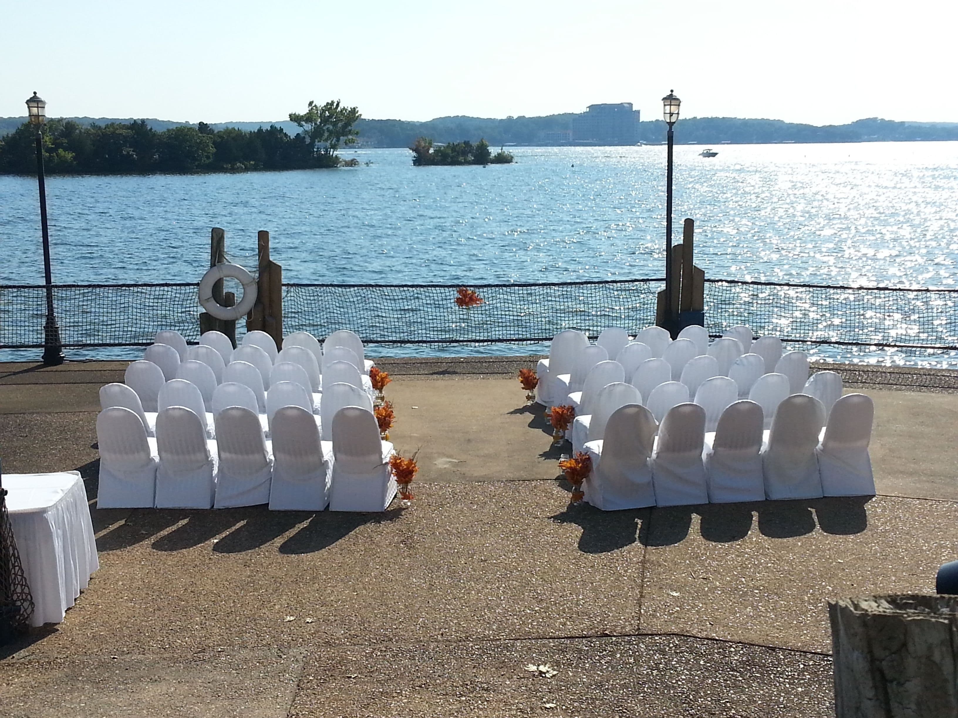 An outdoor seating area with white chairs and tables, a netted barrier, and a view of a body of water with boats and distant buildings.