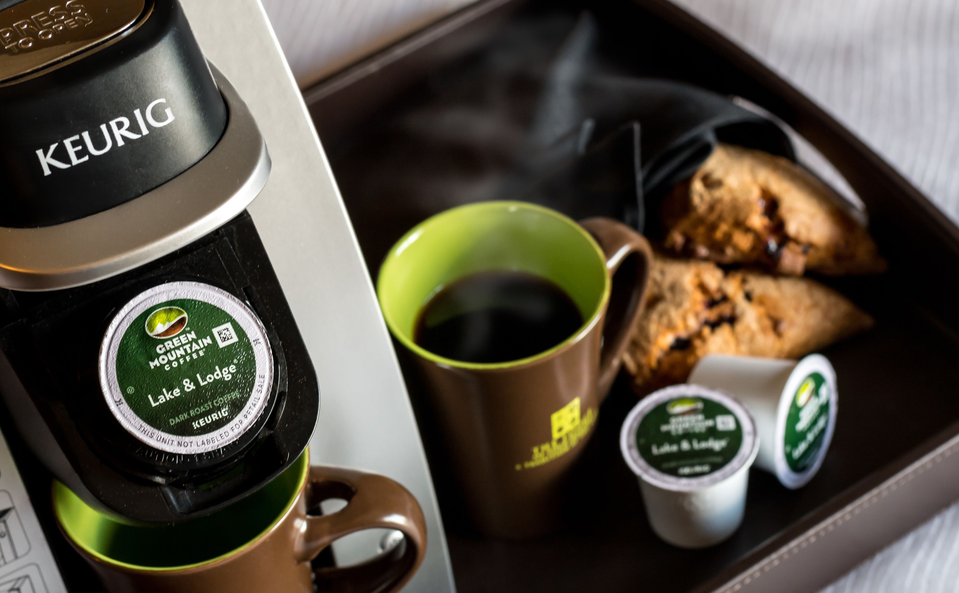 A Keurig coffee maker with a green Mountain Coffee logo, a brown mug, a green mug, and two small containers of coffee.