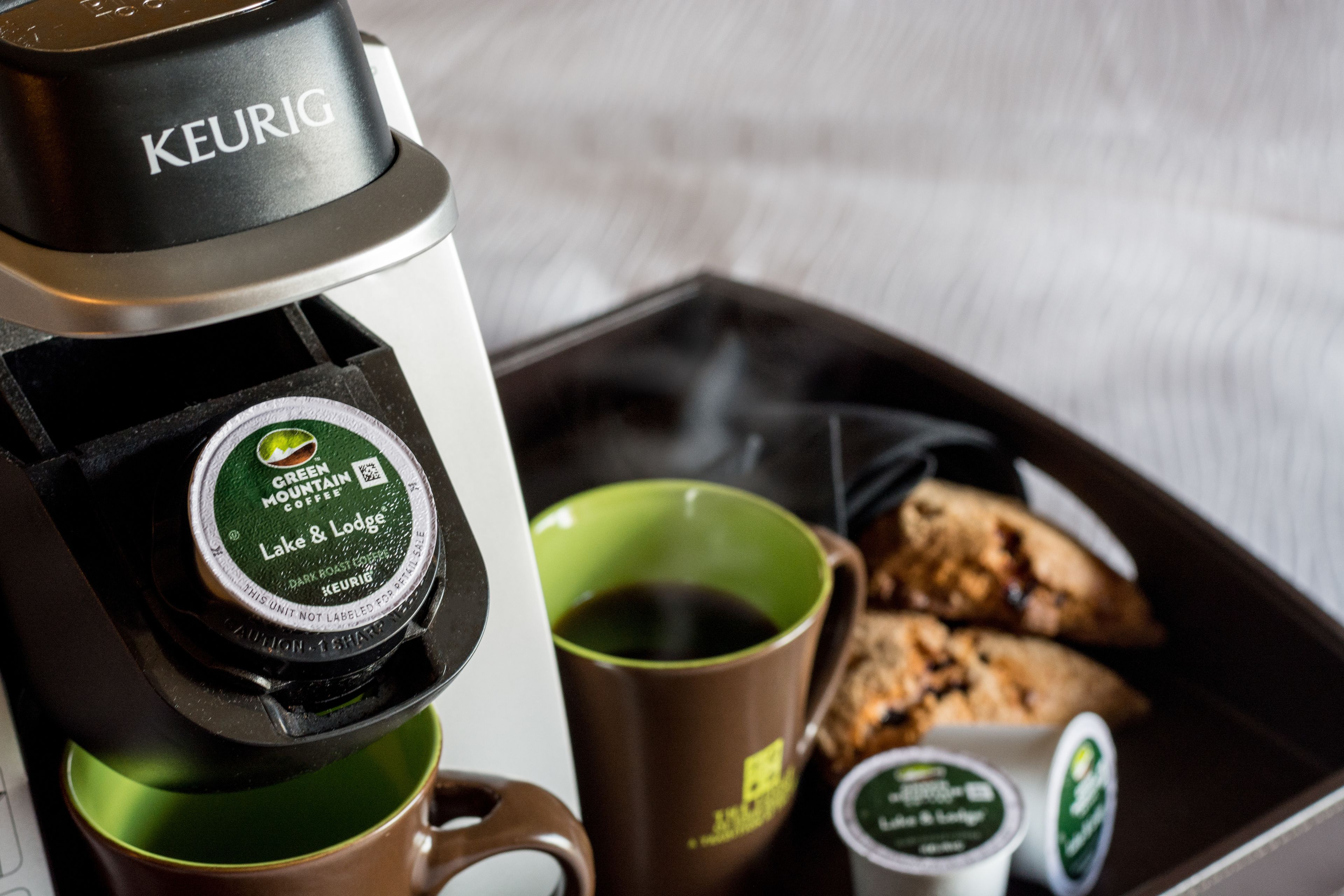 En Keurig-kaffemaskine med en kaffekapsel mærket "Green Mountain Coffee Lake & Lodge", en grøn kop, en brun kop samt en bakke med kager og en lille beholder med fløde.