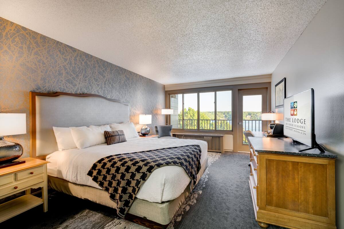 Une chambre d’hôtel avec un grand lit, une table de chevet en bois, un bureau, une commode et une grande fenêtre donnant sur l’extérieur.