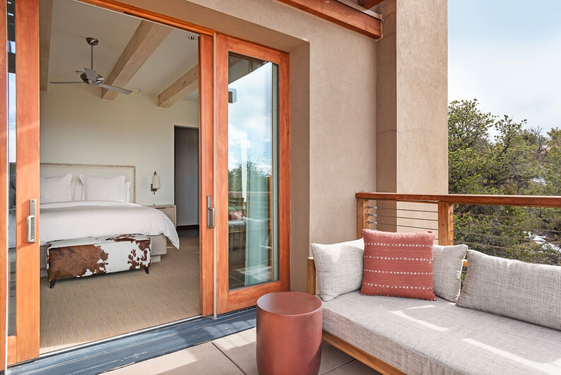 A balcony with a sofa and cushions, a sliding glass door leading to a bedroom with a bed and a nightstand, and a wooden ceiling beam.